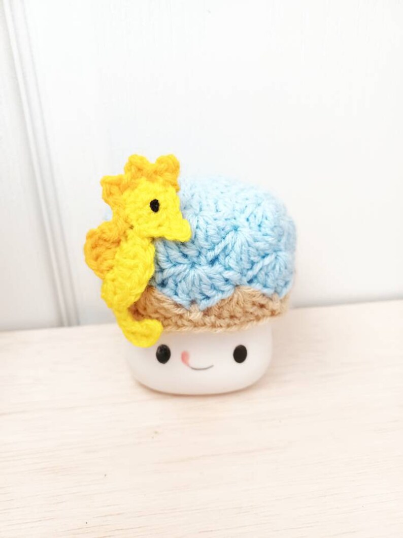 May include: A white ceramic marshmallow with a blue and brown crocheted hat and a yellow crocheted seahorse on top.