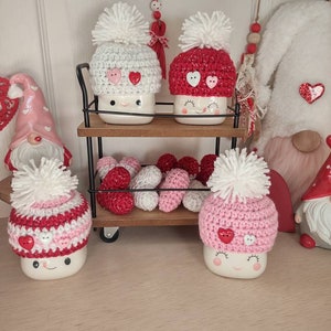 May include: Four white ceramic mugs with pink and white crocheted hats and red hearts. The mugs are on a black metal cart with a wooden shelf. There are pink and white hearts on the shelf.
