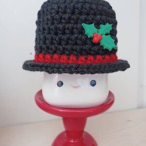 May include: A white ceramic mug with a smiling face, wearing a grey crocheted hat with a red trim and a green holly accent.