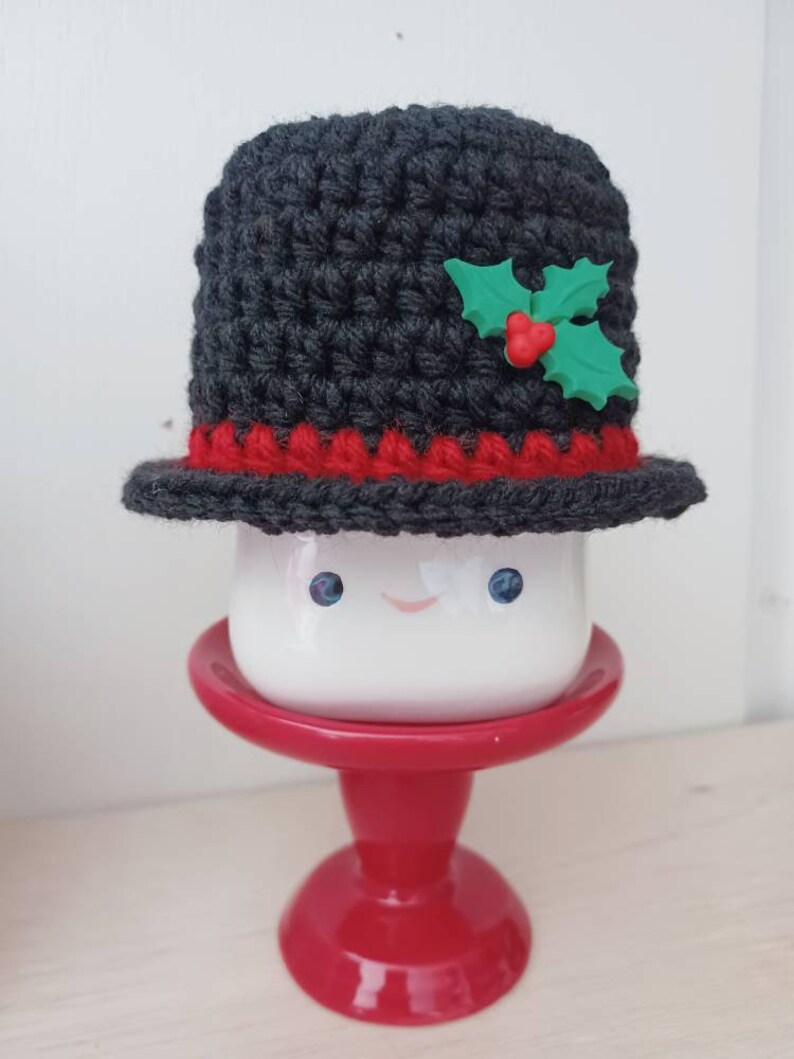 May include: A white ceramic mug with a smiling face, wearing a grey crocheted hat with a red trim and a green holly accent.