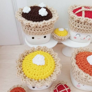 May include: Five crocheted pie mug toppers in various colors and designs. The toppers are made with brown, yellow, orange, and red yarn. The toppers are topped with white yarn to represent whipped cream.