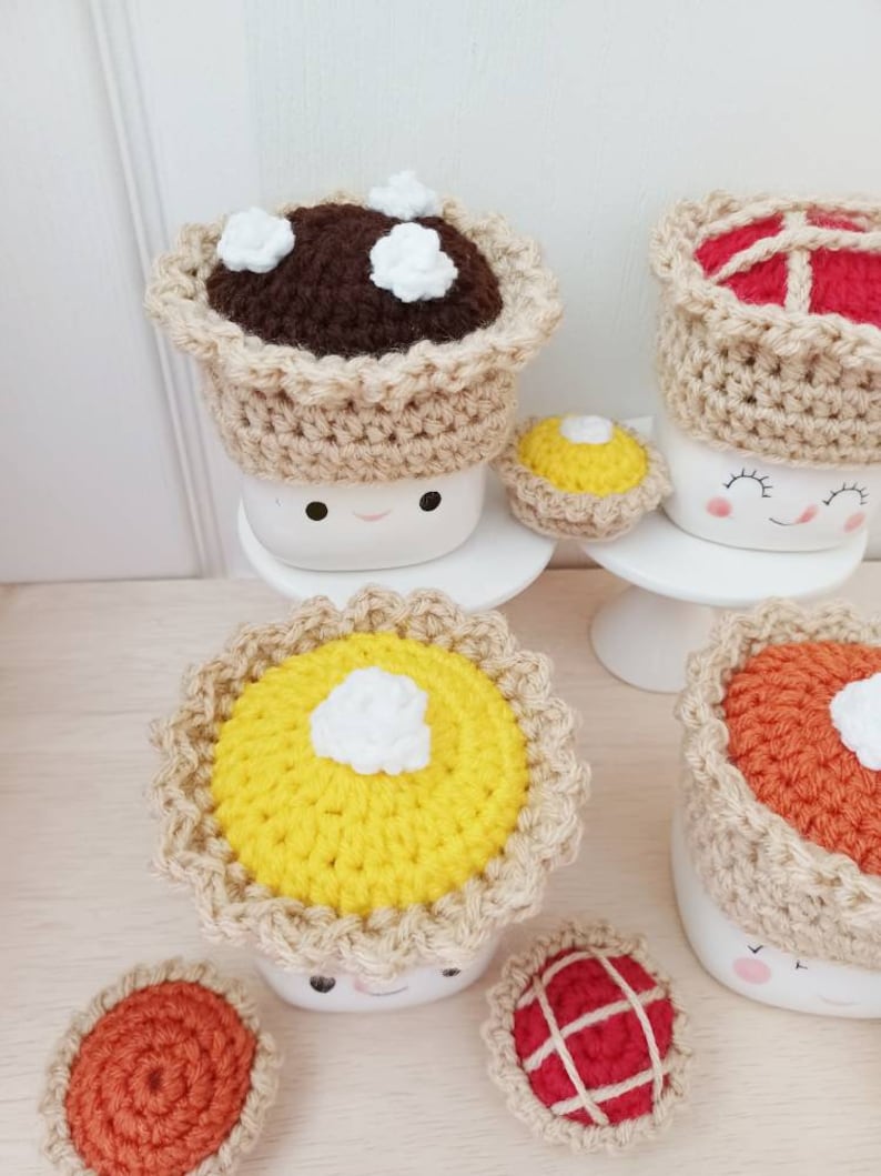 May include: Five crocheted pie mug toppers in various colors and designs. The toppers are made with brown, yellow, orange, and red yarn. The toppers are topped with white yarn to represent whipped cream.