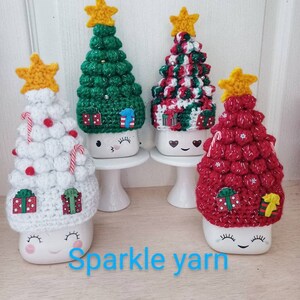 May include: Four white ceramic mugs with crocheted Christmas tree toppers. The trees are green, red, white and red and white. The mugs have a cute face with eyes and a smile. The text "Sparkle yarn" is visible below the mugs.