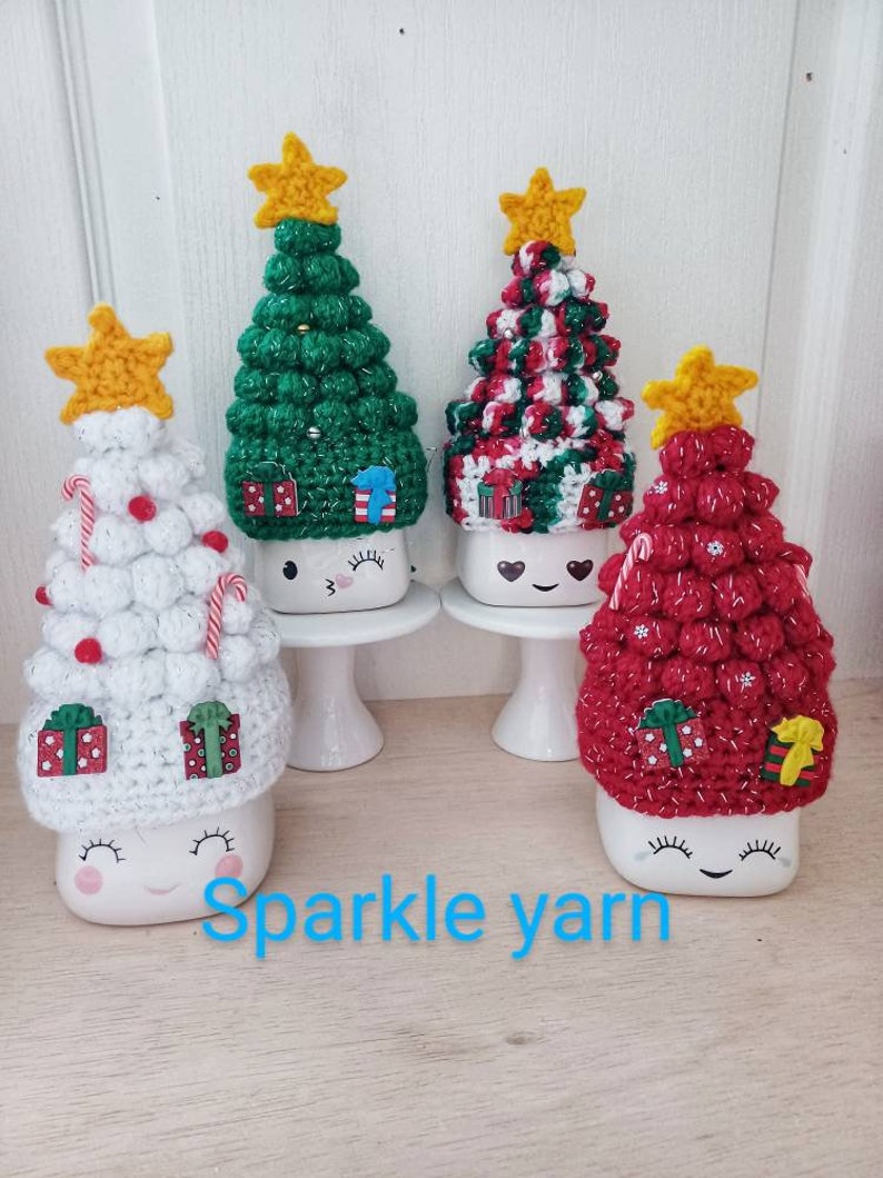 May include: Four white ceramic mugs with crocheted Christmas tree toppers. The trees are green, red, white and red and white. The mugs have a cute face with eyes and a smile. The text "Sparkle yarn" is visible below the mugs.