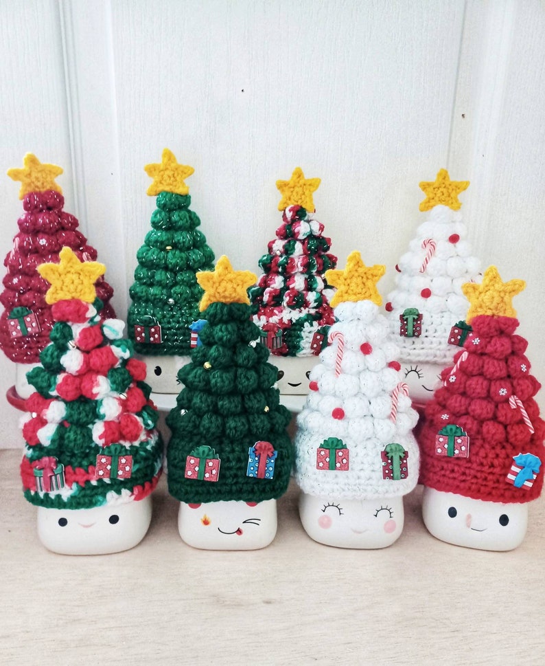 May include: Six white ceramic mugs with crocheted Christmas tree hats. The hats are in various colors and patterns, including red, green, white, and candy cane stripes. Each mug has a smiling face.