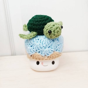May include: A crocheted blue and brown hat with a green and brown crocheted turtle on top. The hat is sitting on a white mug with a smiling face.