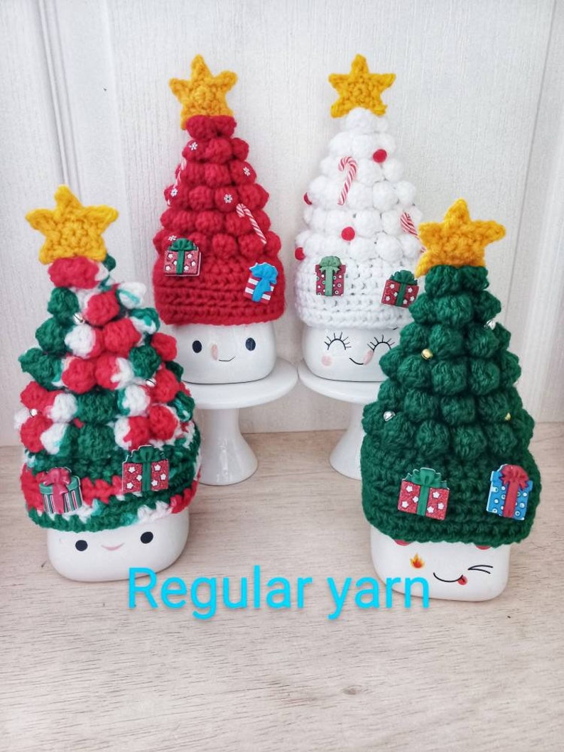 May include: Four crocheted Christmas tree mug toppers in red, white, and green with a yellow star on top. Each topper has a white mug underneath with a happy face.