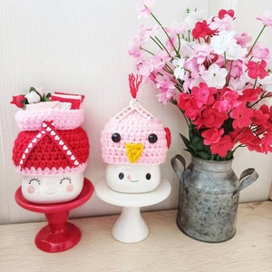 May include: Two white ceramic mugs with crocheted hats. One mug has a pink hat with a yellow beak and the other has a red and white hat with hearts. Both mugs have a smiling face. The mugs are on white pedestals and a red pedestal. A vase with pink and red flowers is in the background.