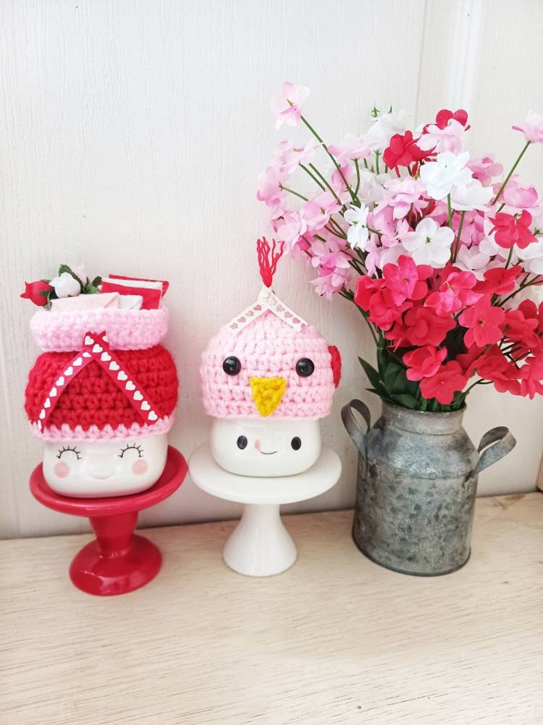 May include: Two white ceramic mugs with crocheted hats. One mug has a pink hat with a yellow beak and the other has a red and white hat with hearts. Both mugs have a smiling face. The mugs are on white pedestals and a red pedestal. A vase with pink and red flowers is in the background.
