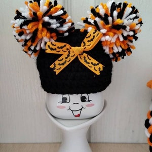 May include: A black crocheted hat with orange and white pom-poms and a black and orange bat ribbon. The hat is sitting on a white ceramic mug with a smiling face.
