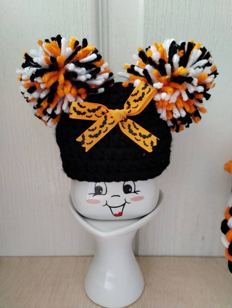 May include: A black crocheted hat with orange and white pom-poms and a black and orange bat ribbon. The hat is sitting on a white ceramic mug with a smiling face.