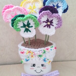May include: A crocheted flower arrangement in a white pot with a smiling face. The flowers are in various colors, including pink, yellow, green, purple, and blue. The pot has a ribbon with a butterfly pattern around it. The arrangement is decorated with a purple ribbon that says "Happy Mother's Day".