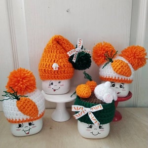 May include: Four white mugs with crocheted hats and carrot designs. The mugs have happy faces and are decorated with orange and green yarn.