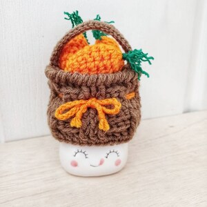 May include: A white ceramic mug with a smiling face, wearing a brown crocheted basket hat with a yellow bow and filled with orange crocheted carrots.