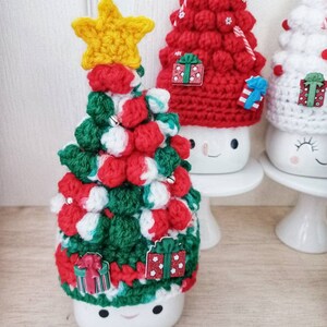 May include: Three white ceramic mugs with crocheted Christmas tree hats. The mugs have painted smiley faces. The trees are decorated with red and white pom-poms, green and red stripes, and small gift ornaments.