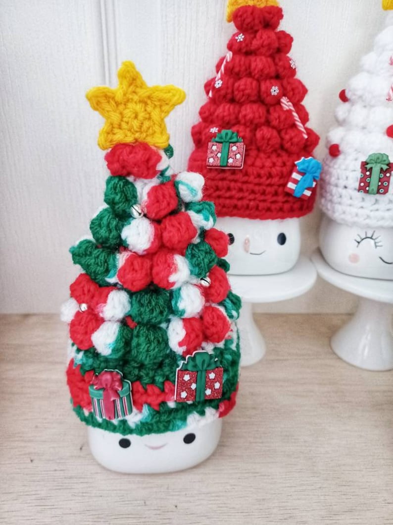 May include: Three white ceramic mugs with crocheted Christmas tree hats. The mugs have painted smiley faces. The trees are decorated with red and white pom-poms, green and red stripes, and small gift ornaments.