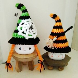 May include: Two white ceramic marshmallows with painted faces wearing crocheted witch hats. The hats are black and green with a white spiderweb design and black and orange with a broom design. The marshmallows are sitting on wooden stools.