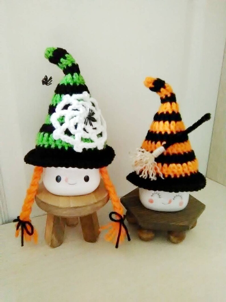 May include: Two white ceramic marshmallows with painted faces wearing crocheted witch hats. The hats are black and green with a white spiderweb design and black and orange with a broom design. The marshmallows are sitting on wooden stools.