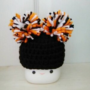 May include: A black crocheted hat with orange, black, and white pom-poms on top. The hat is sitting on a white mug with a smiley face.