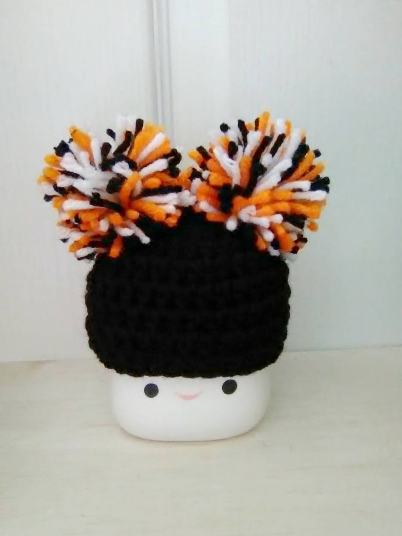 May include: A black crocheted hat with orange, black, and white pom-poms on top. The hat is sitting on a white mug with a smiley face.
