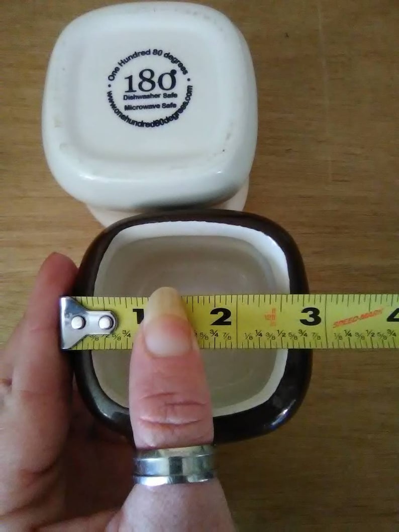 May include: Two square ceramic ramekins, one white with a blue logo that says "One Hundred 80 degrees" and "180" and the other black with a white interior. A yellow measuring tape is wrapped around the black ramekin, measuring 3 inches in diameter.