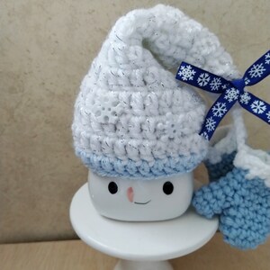 K&ouml;nnte beinhalten: Wei&szlig;er H&auml;kelhut mit blauem Besatz und einem blauen Band mit wei&szlig;en Schneeflocken, auf einer wei&szlig;en Tasse mit einem Schneemann-Gesicht. Die Tasse steht auf einem wei&szlig;en Kuchenst&auml;nder.