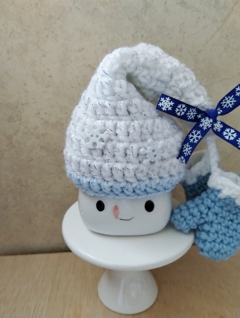 K&ouml;nnte beinhalten: Wei&szlig;er H&auml;kelhut mit blauem Besatz und einem blauen Band mit wei&szlig;en Schneeflocken, auf einer wei&szlig;en Tasse mit einem Schneemann-Gesicht. Die Tasse steht auf einem wei&szlig;en Kuchenst&auml;nder.