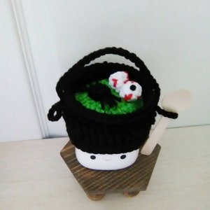 May include: A black crocheted cauldron with green and white googly eyes, sitting on a wooden stand. The cauldron has a wooden spoon.