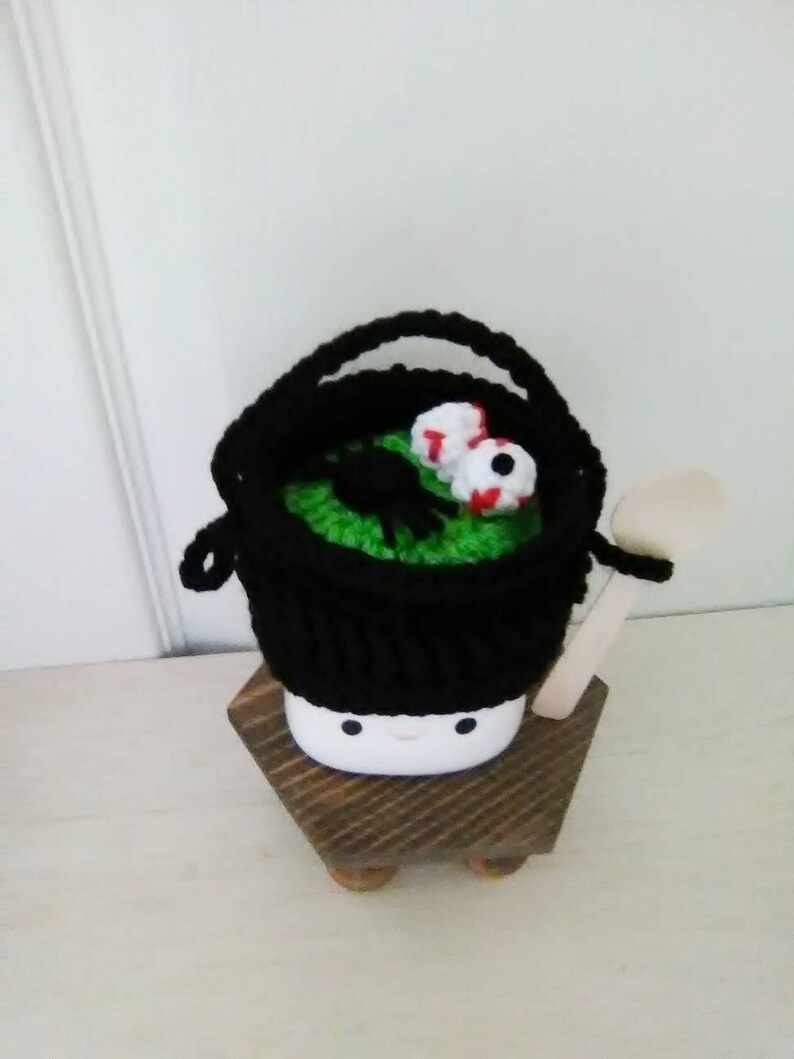 May include: A black crocheted cauldron with green and white googly eyes, sitting on a wooden stand. The cauldron has a wooden spoon.
