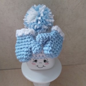 K&ouml;nnte beinhalten: Eine hellblaue Strickm&uuml;tze mit einem wei&szlig;en Bommel und passenden F&auml;ustlingen. Die M&uuml;tze sitzt auf einer wei&szlig;en Keramiktasse mit einem l&auml;chelnden Gesicht.