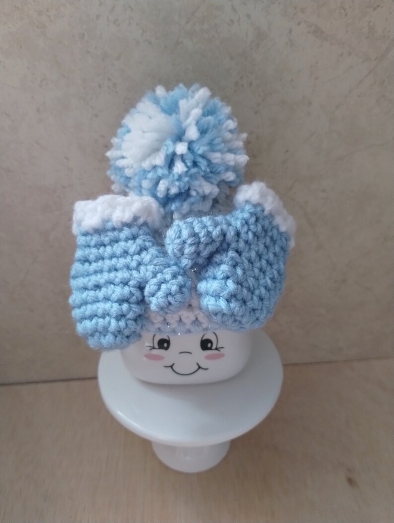 K&ouml;nnte beinhalten: Eine hellblaue Strickm&uuml;tze mit einem wei&szlig;en Bommel und passenden F&auml;ustlingen. Die M&uuml;tze sitzt auf einer wei&szlig;en Keramiktasse mit einem l&auml;chelnden Gesicht.