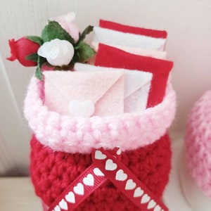 May include: A crocheted red and pink planter with a white ribbon with hearts, filled with pink and red felt envelopes and a small bouquet of pink and red roses.
