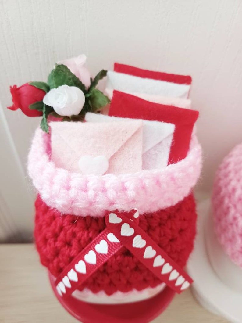 May include: A crocheted red and pink planter with a white ribbon with hearts, filled with pink and red felt envelopes and a small bouquet of pink and red roses.