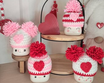 Könnte beinhalten: Vier weiße Keramikbecher mit rosa und roten Häkelmützen. Jede Mütze hat ein pinkfarbenes Herz auf der Vorderseite. Die Becher sind auf einem zweistufigen schwarzen Metallständer angeordnet.