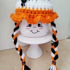 May include: A crocheted Halloween hat with orange, black, and white yarn. The hat has two long braids with black spiders attached. The hat is sitting on a white cake stand.