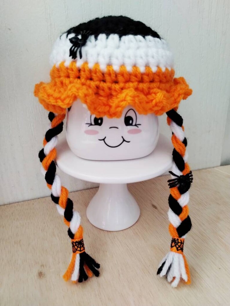 May include: A crocheted Halloween hat with orange, black, and white yarn. The hat has two long braids with black spiders attached. The hat is sitting on a white cake stand.