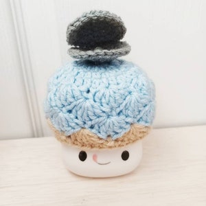 May include: A crocheted blue and tan hat with a grey crocheted clam on top. The hat is on a white ceramic marshmallow with a smiling face.