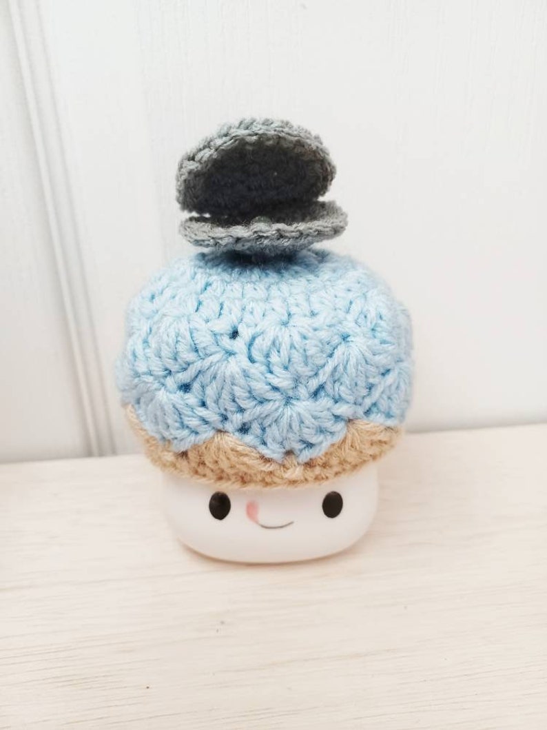 May include: A crocheted blue and tan hat with a grey crocheted clam on top. The hat is on a white ceramic marshmallow with a smiling face.