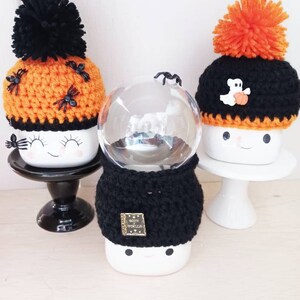 K&ouml;nnte beinhalten: Drei Halloween-Keramikbecher mit geh&auml;kelten H&uuml;ten und Bommeln. Ein Becher hat einen orangefarbenen Hut mit schwarzen Spinnen, ein anderer einen schwarzen Hut mit einem Geist und einem K&uuml;rbis, und der dritte hat eine schwarze H&uuml;lle. Die Becher stehen auf schwarzen und wei&szlig;en St&auml;ndern.