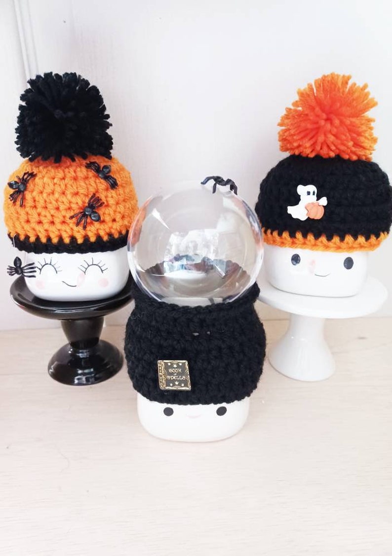 K&ouml;nnte beinhalten: Drei Halloween-Keramikbecher mit geh&auml;kelten H&uuml;ten und Bommeln. Ein Becher hat einen orangefarbenen Hut mit schwarzen Spinnen, ein anderer einen schwarzen Hut mit einem Geist und einem K&uuml;rbis, und der dritte hat eine schwarze H&uuml;lle. Die Becher stehen auf schwarzen und wei&szlig;en St&auml;ndern.