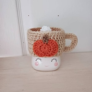 May include: A crocheted mug cozy with a pumpkin design in orange and brown, on a white ceramic mug with a smiling face.