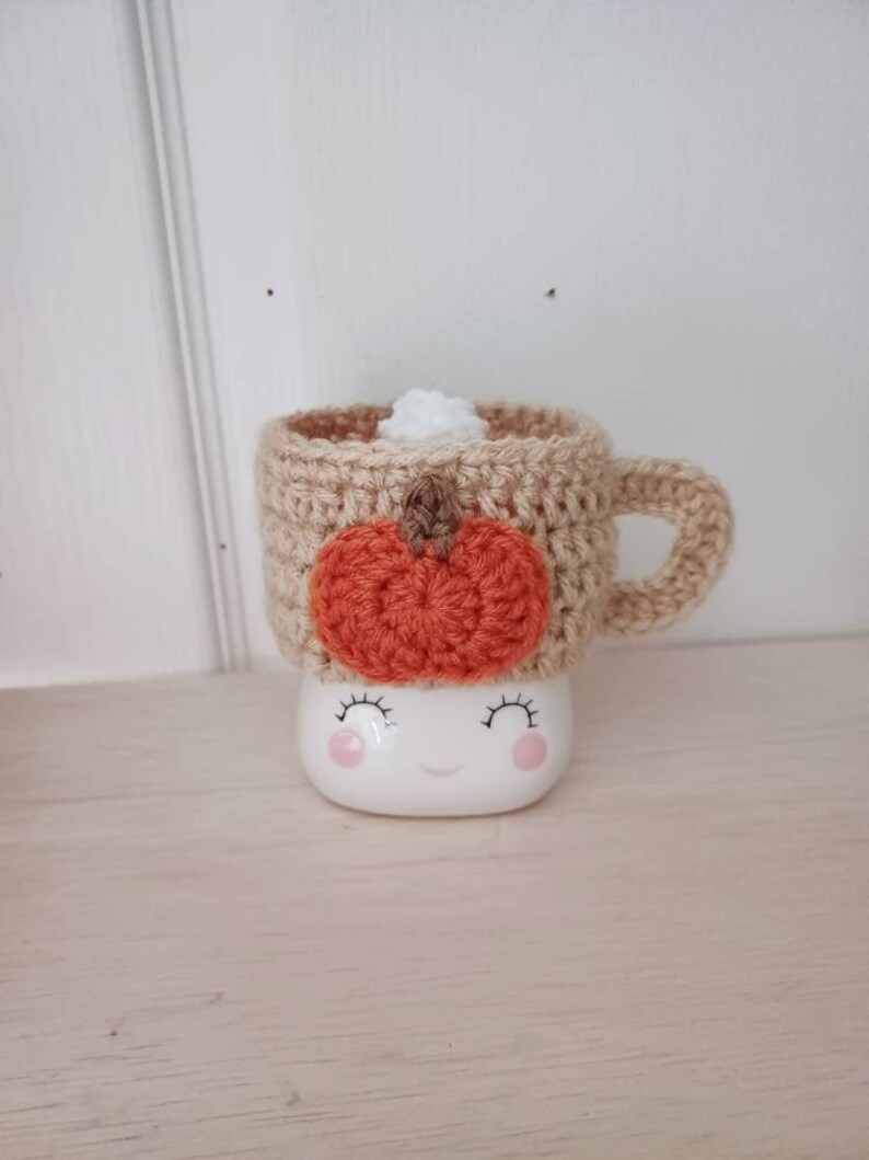 May include: A crocheted mug cozy with a pumpkin design in orange and brown, on a white ceramic mug with a smiling face.
