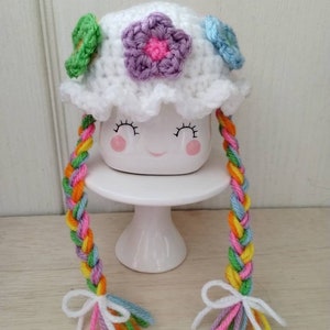 May include: White crocheted hat with colorful flower accents and long braided yarn tails with white bows. The hat is on a white ceramic mug with a smiling face.