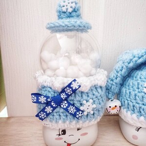 May include: A whimsical snowman-themed snow globe. The globe features a clear, round container filled with white fluff, topped with a light blue crocheted hat with a snowflake accent. The base is white with a winking face and a blue bow.