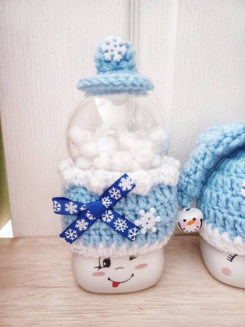 May include: A whimsical snowman-themed snow globe. The globe features a clear, round container filled with white fluff, topped with a light blue crocheted hat with a snowflake accent. The base is white with a winking face and a blue bow.