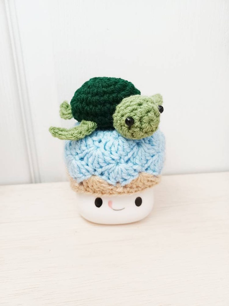 May include: A crocheted blue and brown hat with a green and brown crocheted turtle on top. The hat is sitting on a white mug with a smiling face.