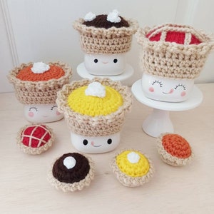 May include: A set of six crocheted mini pies in various colors and fillings, including chocolate, pumpkin, and apple. The pies are topped with white crocheted dollops of whipped cream. The pies are displayed on a white surface.
