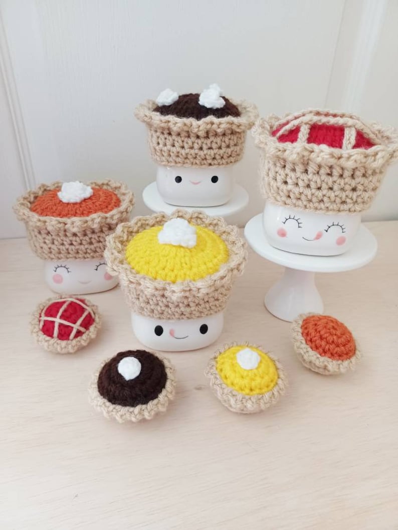 May include: A set of six crocheted mini pies in various colors and fillings, including chocolate, pumpkin, and apple. The pies are topped with white crocheted dollops of whipped cream. The pies are displayed on a white surface.