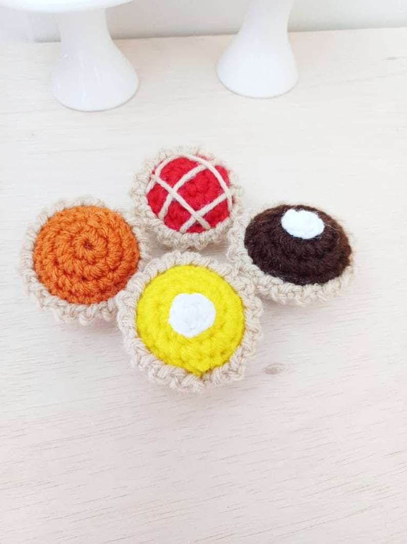 May include: Four crocheted mini pies with different fillings: orange, red, brown, and yellow. Each pie has a white crocheted crust.