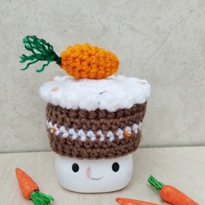 May include: A white ceramic mug with a brown and white crocheted carrot cake hat. The mug has a smiling face and black eyes. There are four small orange crocheted carrots on a wooden surface.